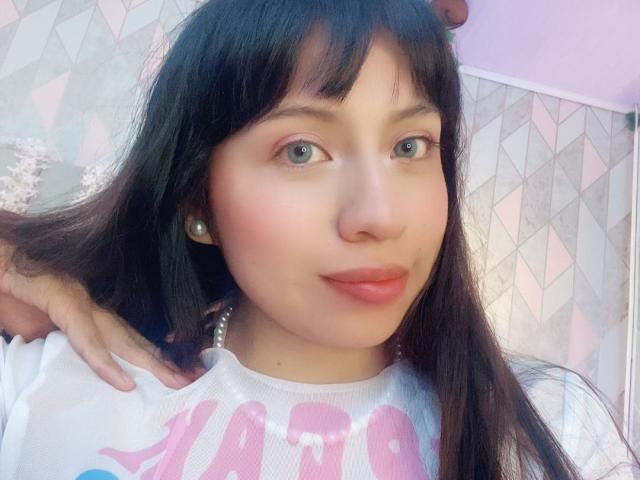 ZoeRedds has long black hair, green eyes, and pink lipstick. She's wearing a white shirt with pink text, and a hand is in her hair. She's looking at the camera.