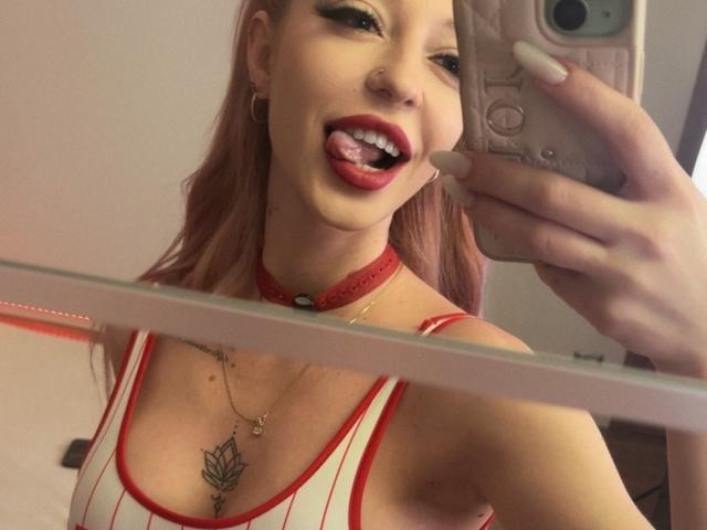 MilanaAmber con i capelli rosa, una collana rossa e una canottiera a righe, che mostra la lingua, con le tette in vista in un selfie allo specchio, che tiene un telefono rosa, con un anello al naso e tatuaggi visibili..