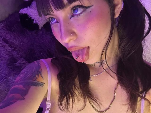 KuroBabee tem pele clara, olhos azuis, rabos de cavalo pretos, um piercing no nariz, um piercing na língua, um braço tatuado e está de língua para fora. Ela está vestindo um sutiã rosa com um colar de corrente..