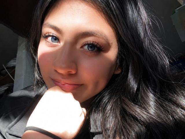 ZoeRedds with shiny black hair, blue eyes, and a cute smile, resting her chin on her hand, sunlight highlighting her face. Her nose is small and her lips are pink.