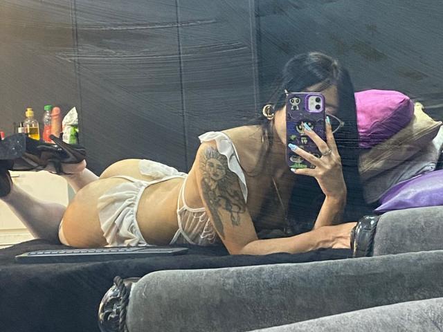 MiaDeLune liegt auf dem Bauch in weißer Unterwäsche, wobei ihr Hintern und ihr tätowierter Arm zu sehen sind. Sie trägt schwarze High Heels und macht ein Selfie mit ihrem Handy, wobei sie sich im Spiegel betrachtet..