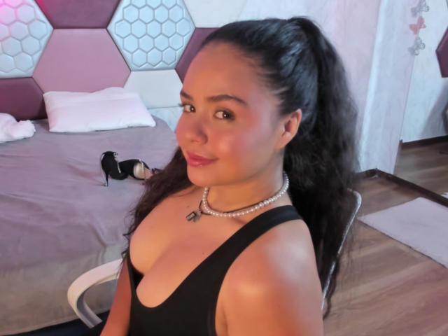 ArielleWilde with long black hair in a ponytail, wearing a black dress with a deep cleavage, pearl necklace, and black high heels, smiling at the camera.