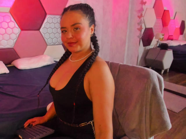 ArielleWilde, with braided hair, black tank top, and chain belt, sits on a chair, showing her tits in a room with pink hexagon-patterned walls. She's smiling.