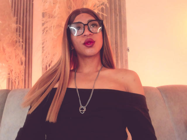 Kalyban with long blonde hair, black off-shoulder top, red lipstick, and glasses, wearing a silver necklace with a heart pendant, sitting on a grey couch.