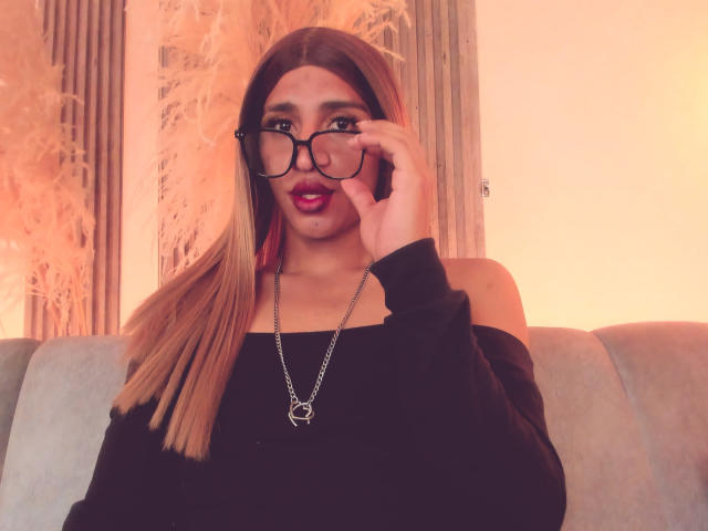 Kalyban with long blonde hair, black glasses, red lipstick, black off-shoulder top, silver necklace, holding glasses, sitting on grey couch, looking at camera.