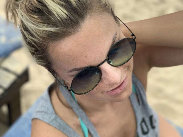 BelleLisaG com cabelo loiro em um coque bagunçado, usando óculos de sol escuros, uma regata cinza e alças de sutiã azuis, sorrindo levemente, mostrando sardas e um nariz fofo. Cenário de praia..
