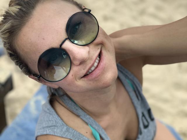 BelleLisaG with blonde hair, wearing round sunglasses and a gray tank top, smiling at the camera, showing a cute, freckled face and a slight tan. Beach setting.
