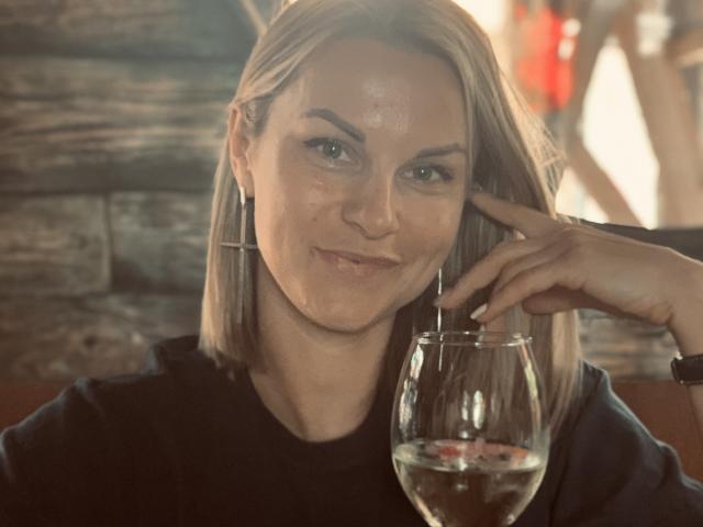 BelleLisaG, avec des cheveux blonds, un haut noir et des boucles d'oreilles, avec un léger sourire, tenant un verre de vin dans sa main droite, son doigt gauche touchant sa joue..