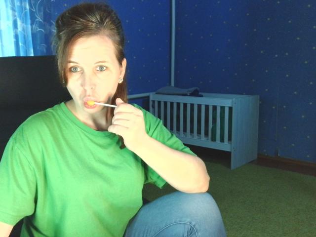 LoveisHt staat met een lolly in haar mond, draagt een groene blouse en blauwe jeans, met haar hand in de buurt van haar mond, en er staat een babybedje op de achtergrond..
