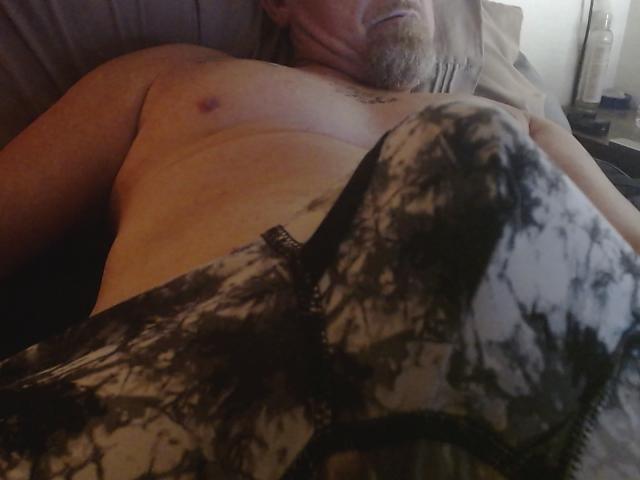 Braadley69 deitado em uma cama, sem camisa, com um cobertor preto e branco estampado cobrindo seus genitais, expondo seu peito peludo e barba. Sua mão está apoiada em seu abdômen..