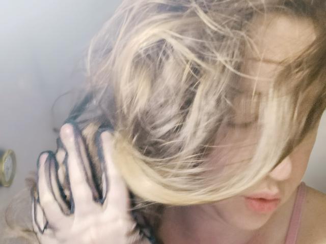SweetLira's blonde hair is being pulled by her hand, covering her face. She has pink lips and is wearing a pink top. Her hand has black and white striped gloves.