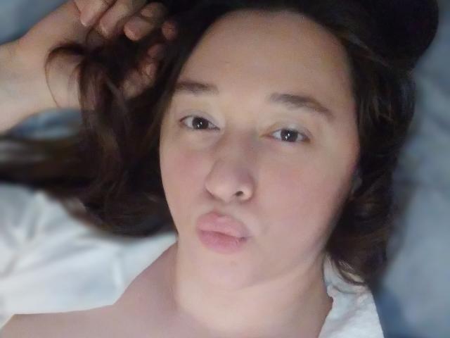 O rosto de SweetLira, com seus olhos castanhos, lábios rosados e finos, pele clara e cabelo castanho escuro, deitada com uma mão no cabelo, vestindo uma camisa branca..