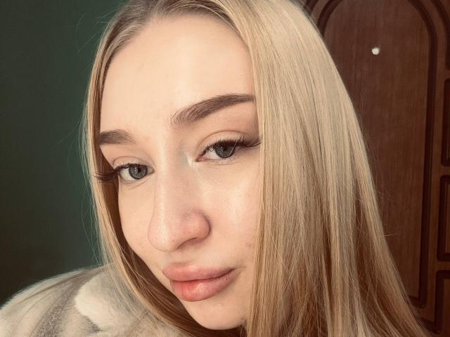 JoetteShigeta's face, blonde hair, blue eyes, full lips, subtle makeup, light skin, thick eyebrows, looking slightly to the side, wearing a beige top, wooden door in background.