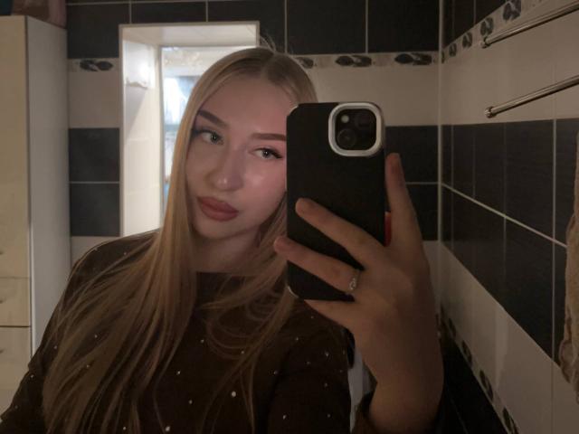 Blonde JoetteShigeta with long hair, pink eyeliner, and red lipstick, holding a black phone, wearing a sheer black top, taking a mirror selfie in a tiled bathroom.