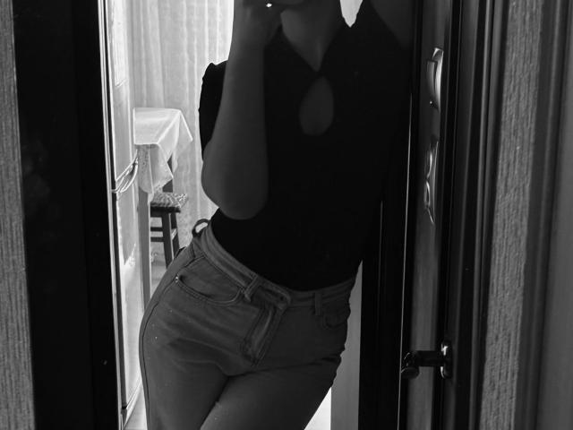 Selfie en blanco y negro de JoetteShigeta, mostrando sus pezones a través de un corte en una camiseta negra ajustada, con jeans de cintura alta, de pie en un marco de puerta, sosteniendo un teléfono..