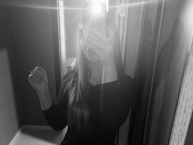Zwart-wit selfie van JoetteShigeta met lang haar, waarbij ze haar gezicht bedekt met een telefoon, gekleed in een donker shirt met lange mouwen, met haar rechtervuist omhoog, heldere flits in de spiegel..