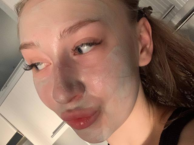 O rosto de Joette Shigeta está coberto por uma máscara facial branca, com lábios vermelhos, olhando para o lado. Cabelo castanho, delineador preto, pele levemente brilhante. Fundo da cozinha desfocado..