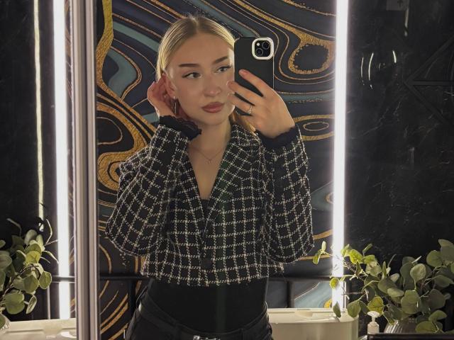 Joette Shigeta macht ein Selfie, trägt ein kariertes Hemd, schwarze, hoch taillierte Hosen, hält ein Telefon, hat blondes Haar, grüne Augen und ein leichtes Lächeln, im gut beleuchteten Badezimmer..