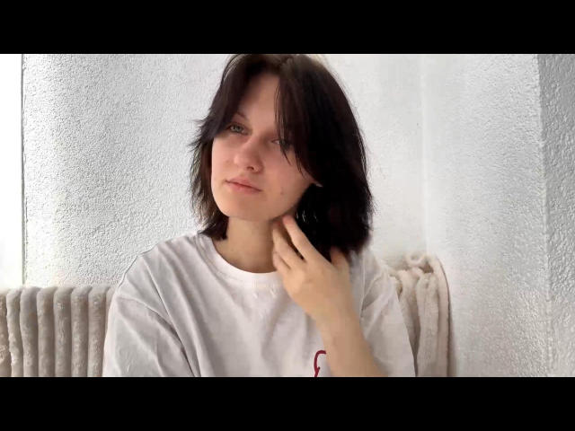 Ada Schorn, com cabelo escuro e desalinhado, vestindo uma camisa branca, tocando na orelha esquerda, com uma expressão ligeiramente confusa, em frente a uma parede branca com textura, com um radiador ao fundo..