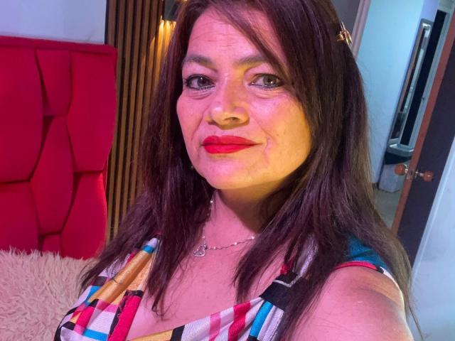 HannaMilff, avec ses cheveux brun moyen, son rouge à lèvres rouge et son haut coloré à motifs, sourit à l'appareil photo. Ses seins sont légèrement visibles sous le haut. Elle porte un collier..