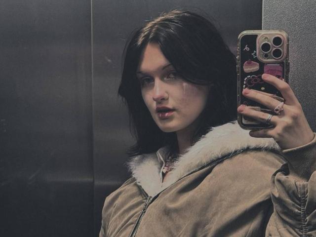 Ada Schorn, con i capelli scuri, indossa una giacca beige con un colletto di pelliccia bianca, e sta scattando un selfie allo specchio con il telefono, mostrando le sue labbra piene e le dita adornate da anelli..