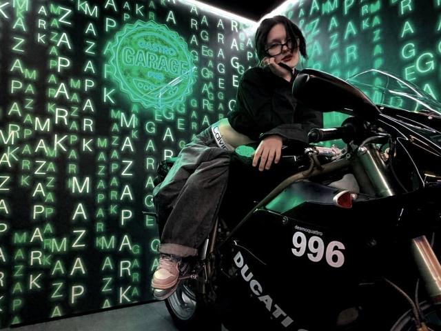 Ada Schorn, avec des lunettes et des cheveux noirs, est appuyée contre une moto Ducati 996, vêtue d'une veste noire, d'un pantalon gris et de baskets roses, dans un garage éclairé d'une lumière vert néon..