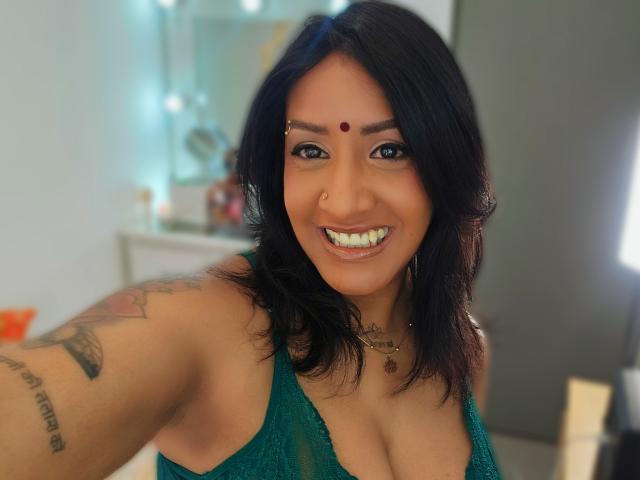 Nahirr has dark hair, a red bindi on her forehead, and is smiling with white teeth. She's wearing a green lace top, showing her tits, with tattoos on her arm.