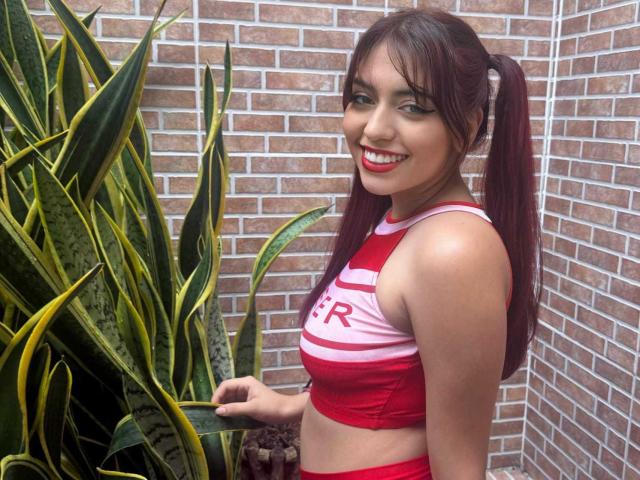 Brendaa Pierce mit roten Zöpfen, die ein rot-pink gestreiftes Crop-Top trägt, lächelt in die Kamera, zeigt ihre Brüste und steht neben grünen und gelben Pflanzen, die wie Schlangen aussehen..