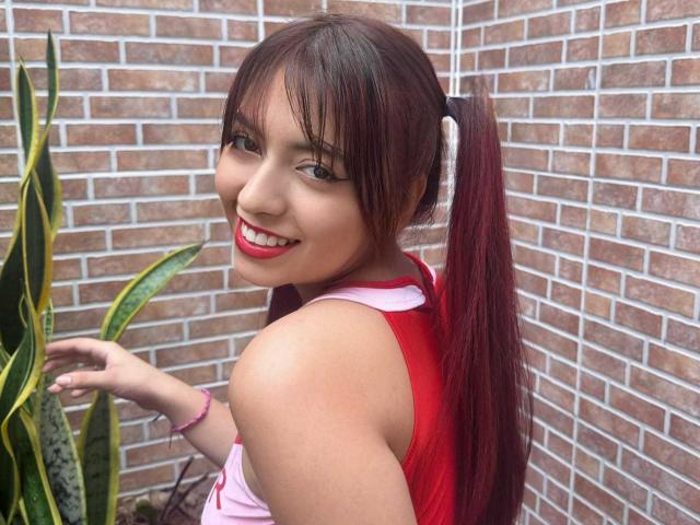 BrendaaPierce with red pigtails, smiling, showing off her tits in a red tank top, pink wristband, standing next to a snake plant, against a brick wall.