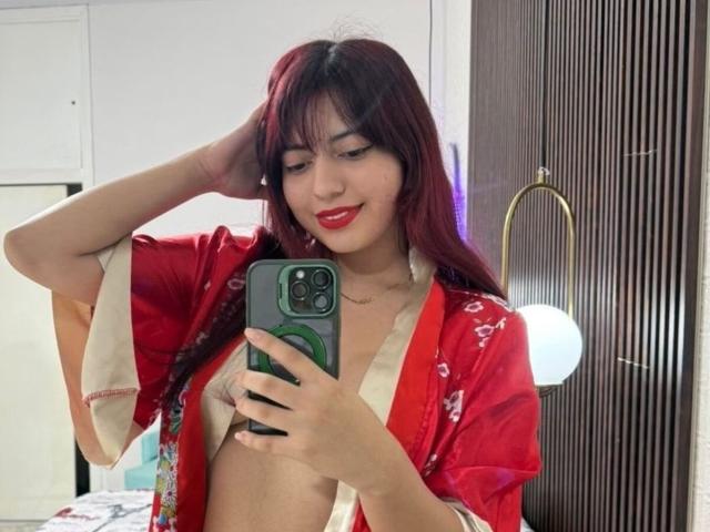 Brendaa Pierce, con i capelli rossi, un kimono floreale rosso e rossetto rosso, tiene in mano un telefono verde, mostrando il suo seno piccolo, è in piedi in una stanza, sorride e ha una mano tra i capelli..
