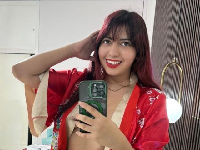 BrendaaPierce with red lipstick, smiling, wearing a red floral kimono, holding a green phone, hand in hair, medium tits, dark red hair, in a well-lit room.