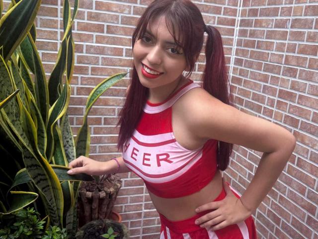 BrendaaPierce with red pigtails, smiling, wearing a red and white cheerleader outfit, pointing at a green snake plant, showing her red cheerleader top and matching skirt.