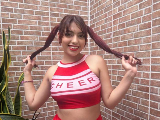 Brendaa Pierce con due codini, maglietta da cheerleader rossa che mostra il seno, sorridente, che tiene i codini, sfondo con un muro di mattoni, braccialetto rosa, rossetto rosso, pianta verde a sinistra..