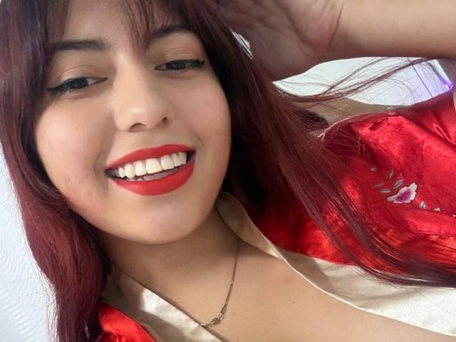 Brendaa Pierce, con lápiz labial rojo, sonriendo y mostrando sus dientes blancos, vestida con una bata de satén roja con bordados florales y un collar de oro, con una mano en el pelo..