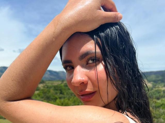 HanahCooper with wet black hair, raising her arm to touch her head, showing off her face with subtle makeup and a slight smile, her tits slightly visible in the background.