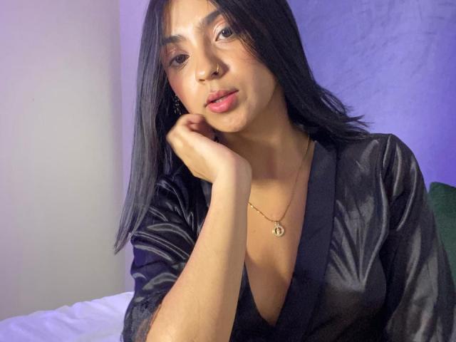 LiliRhodemml, com pele marrom média, cabelo preto e liso, vestindo um robe de cetim preto, colar de ouro, com o queixo apoiado na mão, batom rosa, olhando para a câmera, luz roxa suave..