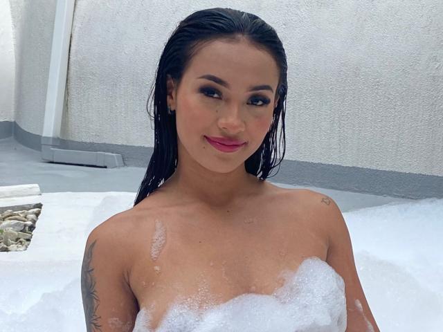 SashaHans with wet black hair, smiling, in a bubbly white bath, showing her tits covered in foam, tattoos on her arm, and pink lipstick.