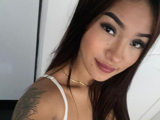 SashaHans with long dark hair, thick eyebrows, and a tattoo on her left shoulder, wearing a white top and gold necklace, smiling with pink lipstick.