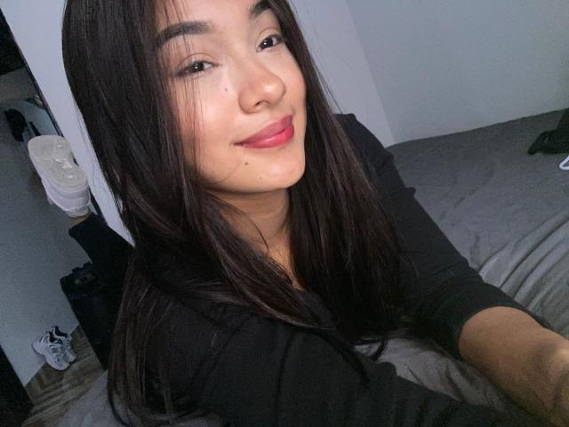 SashaHans has long black hair, a cute smile with pink lips, and is wearing a black shirt, sitting on a bed, showing a small beauty mark on her cheek.
