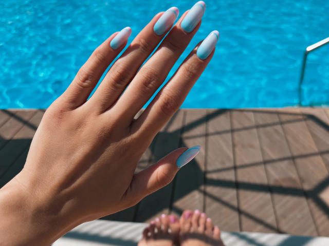 Pinglis Hand mit hellblau lackierten, langen, mandelförmigen Nägeln, die ihre glatte, gebräunte Haut zeigen, befindet sich im Vordergrund. Ihre Zehen mit rosa Nagellack sind im Hintergrund zu sehen..