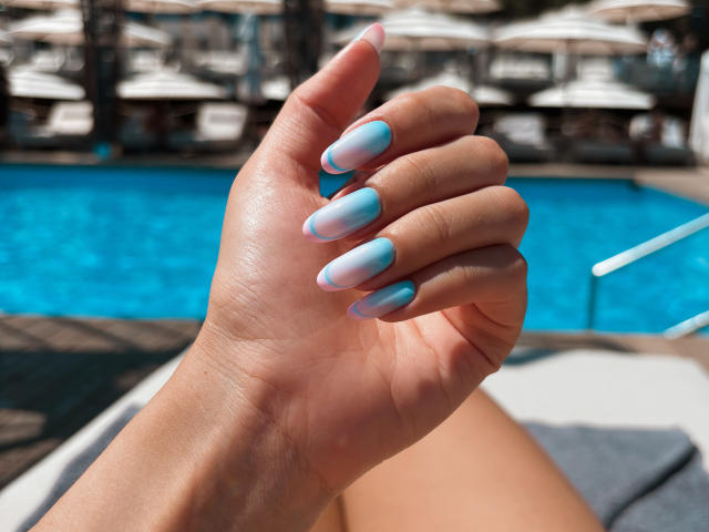 Pingli's hand with pastel blue and pink ombre nails, showing off her long, glossy nails, by a sunlit pool. Her skin is tan and smooth.