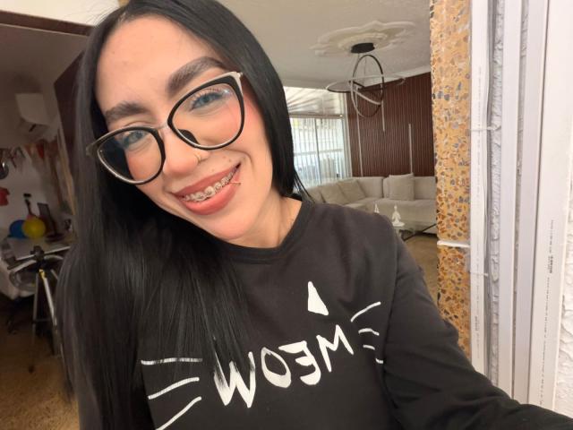 CamilleJuicyHot with black hair, glasses, and a nose ring, smiling with a tongue piercing, wearing a black "WOMEN" sweatshirt, in a cozy room.