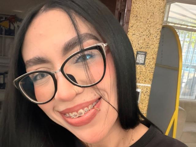 CamilleJuicyHot with black hair, blue eyes, and glasses, showing a metal grin with a lip piercing, smiling at the camera. Her nose is slightly visible.