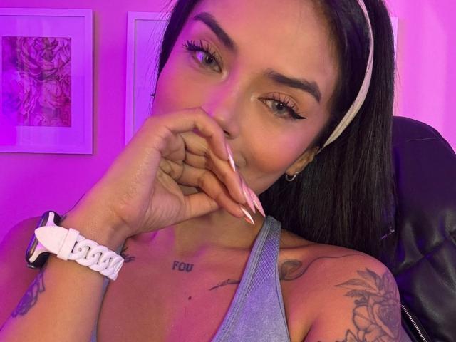 MajoJones with long black hair, white headband, and gray tank top, touching nose with long pink nails, tattoos on chest and arm, pink eyeliner, purple lighting, white scrunchie.