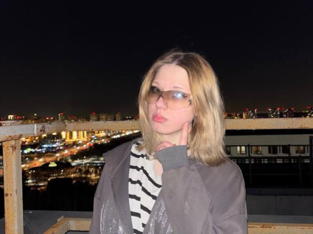 Tamesha Schrimpf, com cabelo loiro, usando óculos e um casaco cinza sobre uma camisa listrada, fazendo beicinho com a mão no pescoço, com as luzes da cidade à noite ao fundo..