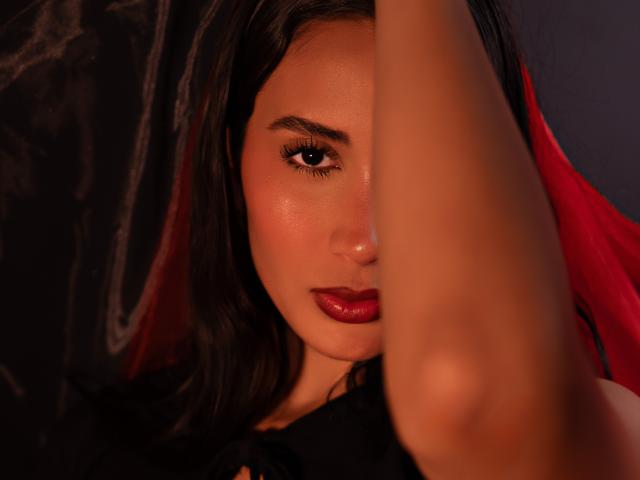 JennTylor's face with dark red lipstick, half-closed eyes, and raised arm in the foreground. Her skin is smooth with a slight sheen. She has black hair and red highlights.