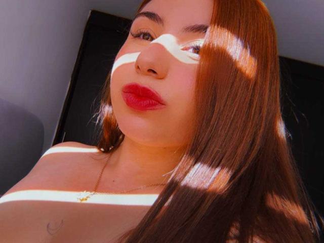 VenusxMusa with red lipstick, long brown hair, and sunlight shadows on her face, wearing a gold necklace, showing her tits slightly in the lower part of the image.