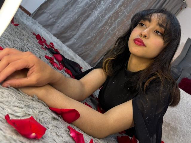 CamiVargas with black hair, red lipstick, and black dress, lying on gray furry bed, surrounded by red rose petals, looking up with a slight pout. Hands resting in front.