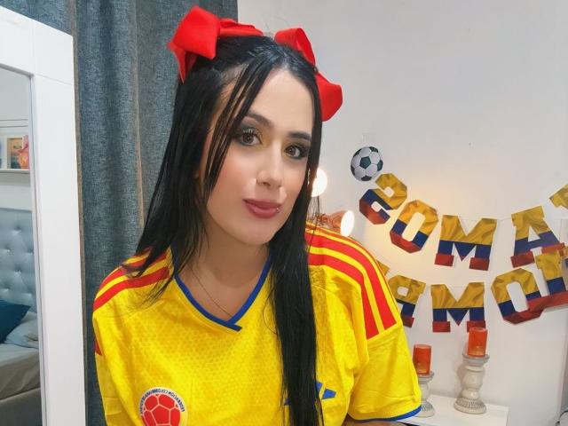 LaurenceRose met lang zwart haar, rode strikken, een geel voetbalsok, rode en gele strepen, een voetbal aan de muur, een witte kamer, blauwe gordijnen en een rode kaars..