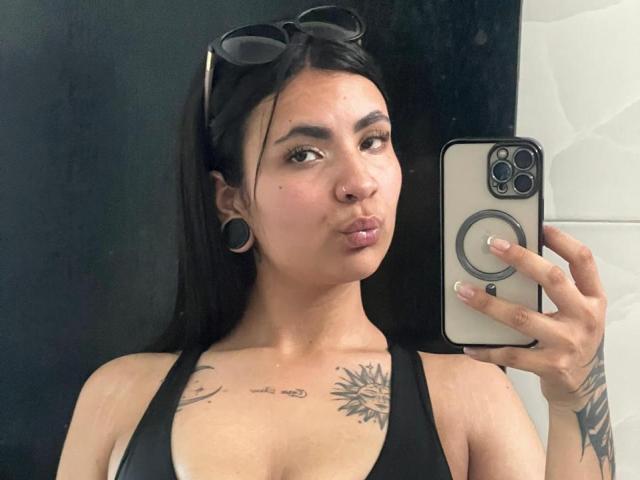 HanahCooper with black hair, wearing sunglasses on head, black tank top, tattoos on chest and arm, taking selfie with iPhone, pouty lips, ear gauges, brown eyes, holding phone.